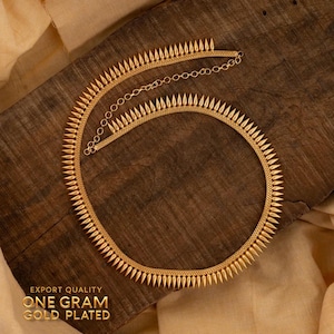 Puede incluir: Un collar dorado con un diseño puntiagudo, sobre una superficie de madera. El collar tiene una cadena fina y está etiquetado como "EXPORT QUALITY ONE GRAM GOLD PLATED". Es una pieza de joyería.