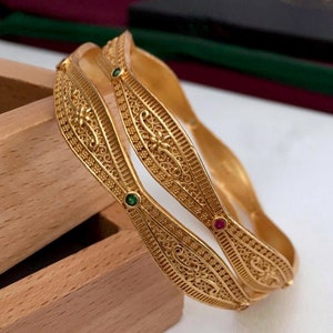 Könnte beinhalten: Zwei goldene Armreifen mit filigranen Designs und kleinen grünen und roten Edelsteinen.