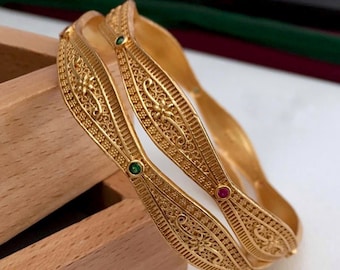 May include: Two gold bangles with intricate designs and small green and red gemstones.