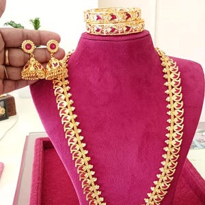 May include: A gold-colored jewelry set featuring a necklace, earrings, and bangles. The necklace and bangles have a repeating design with red accents. The earrings are bell-shaped with red centers. The jewelry is displayed on a red velvet bust.