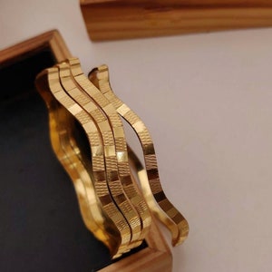 Könnte beinhalten: Satz von vier goldfarbenen Armreifen mit einem wellenförmigen, strukturierten Design. Die Armreifen sind übereinander in einer Holzkiste gestapelt.