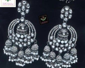 May include: A pair of silver dangle earrings with a large round design and multiple tiers of smaller round ornaments. The earrings are adorned with clear stones and white pearls. The text "Uncut Kumdan weight Jhumka Premium Quality" is visible below the earrings.