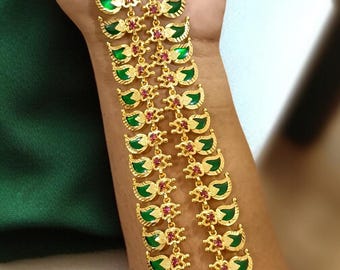 May include: Gold-toned necklace with green and red accents. The necklace features a repeating pattern of green leaf-shaped elements and small red floral designs. A large pendant with a green center and red surrounding stones is at the bottom.