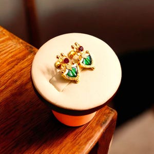 以下が含まれることがあります： シールド型のデザインの金色のイヤリングのペア。各イヤリングは、緑のエナメルセンター、赤い宝石のアクセント、そして精巧なディテールが特徴です。イヤリングはクリーム色のスタンドに展示されています。