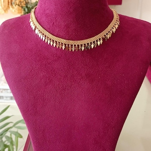 Puede incluir: Un collar dorado con un diseño delicado. El collar presenta una cadena con pequeños colgantes en forma de lágrima. El collar se exhibe en un maniquí granate.