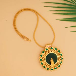 Könnte beinhalten: Goldfarbene Anhängerhalskette mit einem kreisförmigen Anhänger mit grünem und goldenem Design. Der Anhänger hat eine zentrale Figur und rote und grüne Akzente. Die Halskette wird vor einem neutralen Hintergrund präsentiert.