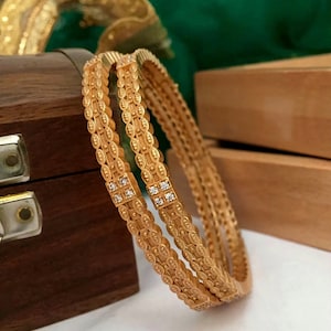 Könnte beinhalten: Zwei goldene Armreifen mit komplizierten Designs und klaren Zirkonia-Steinen. Die Armreifen sind übereinander gestapelt und liegen auf einer Holzoberfläche.