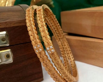 May include: Two gold bangles with intricate designs and clear cubic zirconia stones. The bangles are stacked on top of each other and are resting on a wooden surface.