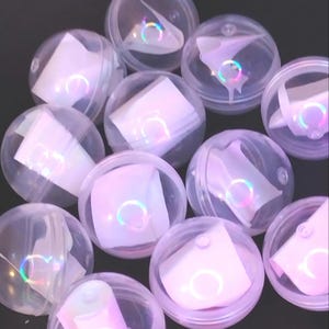 May include: A group of clear plastic spheres containing small, folded pieces of paper with iridescent holographic designs. The spheres are arranged in a random pattern on a dark surface.