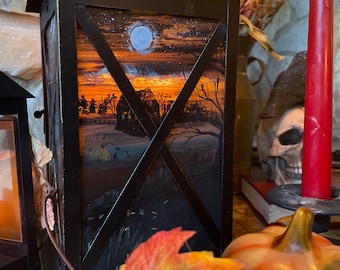 May include: Black metal lantern featuring a painted sunset scene over a rural landscape. The lantern has a decorative top and a glass panel with an X-shaped overlay. A red candle and a decorative pumpkin are in the background.