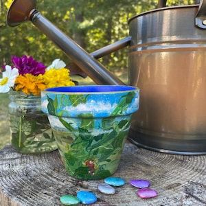Peut inclure: Un pot de fleurs peint à la main avec un ciel bleu et un motif de feuillage vert. Le pot est posé sur une souche d'arbre avec des pierres colorées. Un bocal en verre avec des fleurs et un arrosoir en cuivre sont en arrière-plan.