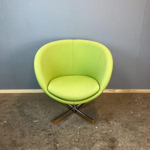 May include: A lime green swivel chair with a rounded back and seat. The chair has a chrome base with wooden accents. The chair is set against a gray wall and a concrete floor.