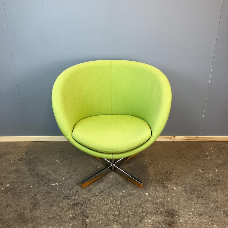May include: A lime green swivel chair with a rounded back and seat. The chair has a chrome base with wooden accents. The chair is set against a gray wall and a concrete floor.