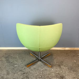 May include: A lime green, egg-shaped swivel chair with a chrome base and wooden accents. The chair's rounded back and seat are upholstered in a solid, light green fabric. The base has a star-like design.
