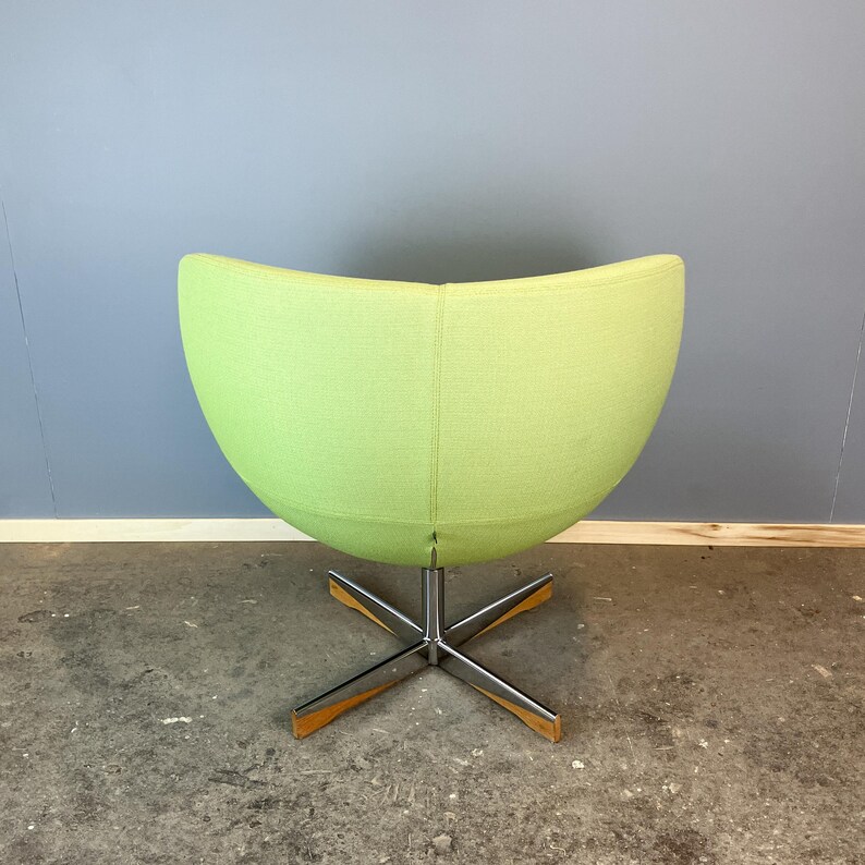 May include: A lime green, egg-shaped swivel chair with a chrome base and wooden accents. The chair's rounded back and seat are upholstered in a solid, light green fabric. The base has a star-like design.