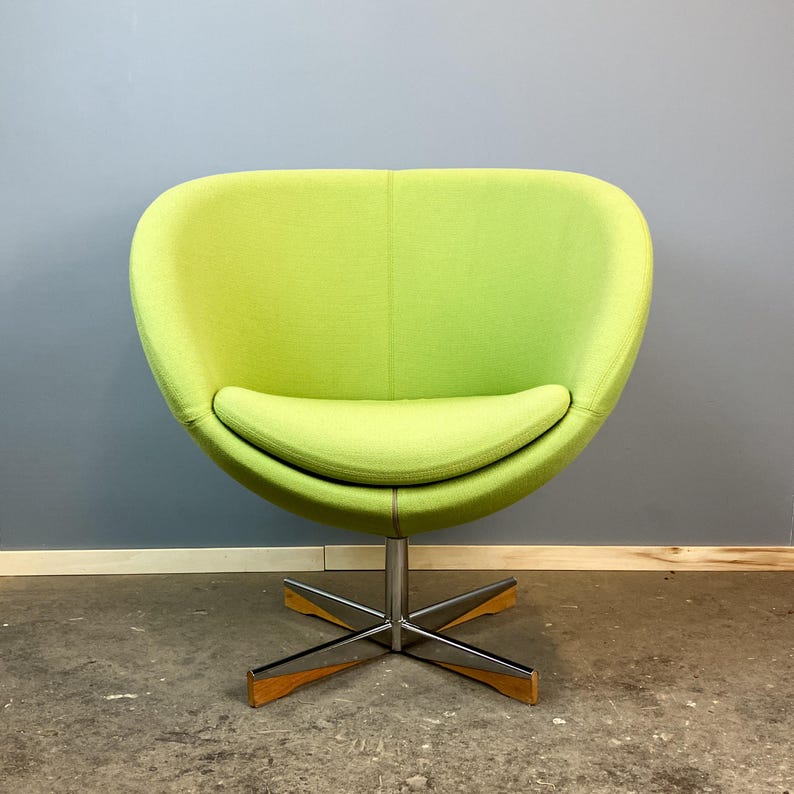 May include: A lime green, mid-century modern style chair. The chair has a rounded back and seat, with a chrome base and wooden accents. The chair is upholstered in a textured fabric and is set against a gray wall.
