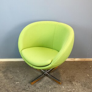 May include: A lime green swivel chair with a rounded back and seat. The chair has a chrome base with wooden accents on the feet. The chair is set against a blue and gray wall.