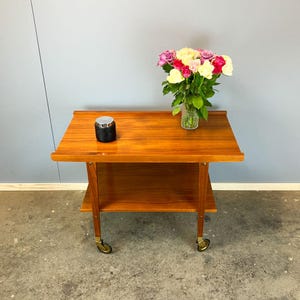 Könnte beinhalten: Ein Mid-Century-Modern-Servierwagen aus Holz mit einem unteren Regal und Messingrollen. Ein kleiner schwarzer Behälter mit silbernem Deckel steht auf der Oberseite. Eine Vase mit bunten Blumen.