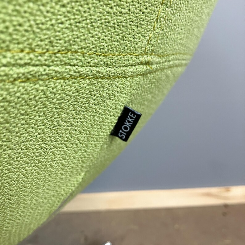 May include: Close-up of a section of a light green upholstered surface. The fabric has a textured appearance with visible stitching. A small black label with the white text "STOKKE" is attached to the fabric.