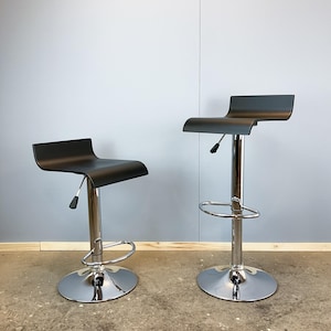 Könnte beinhalten: Zwei schwarze Barhocker mit verchromten Basen und höhenverstellbaren Mechanismen. Die Hocker haben ein geschwungenes Sitzdesign und eine Fußstütze. Die Basen sind rund und reflektierend. Der Hintergrund ist hellgrau.