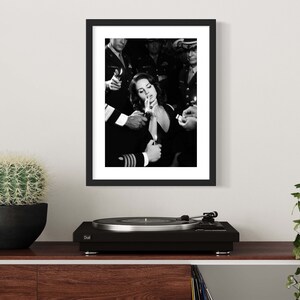 May include: Black and white framed print depicting a woman surrounded by people holding lighters. The image is a vintage-style photograph. The print is displayed on a wooden shelf with a cactus, a record player, and a plant.