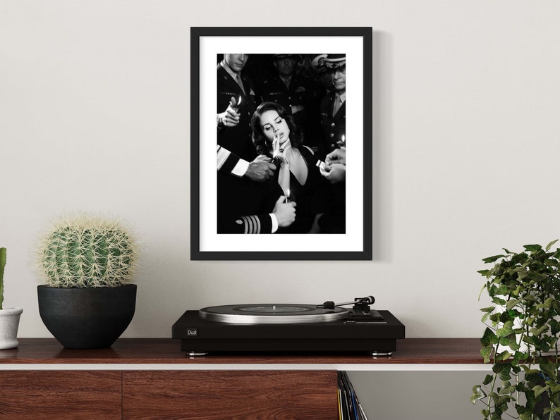 May include: Black and white framed print depicting a woman surrounded by people holding lighters. The image is a vintage-style photograph. The print is displayed on a wooden shelf with a cactus, a record player, and a plant.