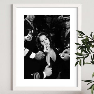 May include: Black and white framed print featuring a woman smoking a cigarette, surrounded by uniformed figures holding lighters. The image has a vintage aesthetic, with a focus on the central figure and the act of smoking.