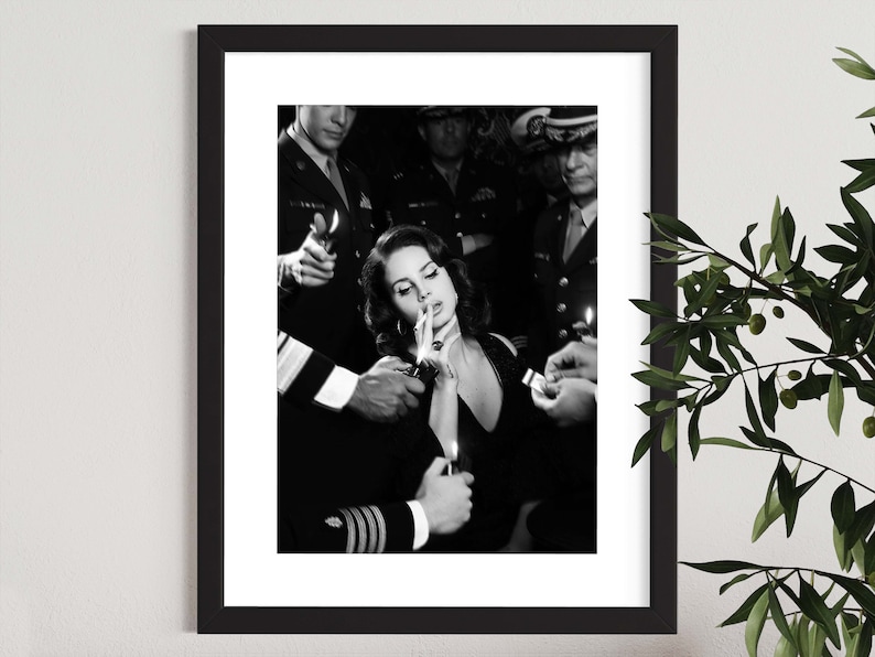 May include: Black and white framed print featuring a woman smoking a cigarette, surrounded by uniformed figures holding lighters. The image has a vintage aesthetic, with a focus on the central figure and the surrounding details.