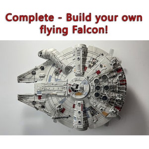 Può includere: Un modello dettagliato dell'astronave Millennium Falcon, con un design complesso di pannelli, tubi e componenti circolari. Il modello è principalmente bianco con accenti grigi e rossi. Il testo in alto recita: "Complete - Build your own flying Falcon!"
