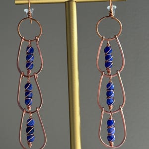 May include: A pair of long dangle earrings featuring three interconnected copper wire teardrop shapes. Each teardrop frame contains a vertical row of two small, round, deep blue beads wrapped in thin copper wire, suspended from a copper hook.