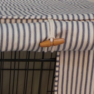 K&ouml;nnte beinhalten: Hundek&auml;figabdeckung aus cremefarbenem Stoff mit vertikalen marineblauen Streifen. Sie hat einen Holzschlie&szlig;enverschluss. Die Abdeckung ist f&uuml;r einen Drahtk&auml;fig konzipiert. Der Stoff scheint ein weiches Baumwollmaterial zu sein.