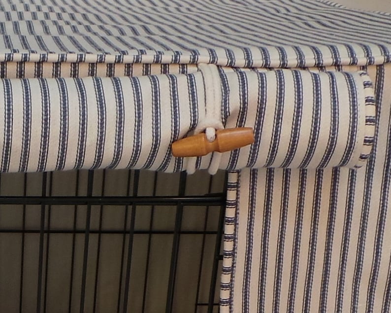 K&ouml;nnte beinhalten: Hundek&auml;figabdeckung aus cremefarbenem Stoff mit vertikalen marineblauen Streifen. Sie hat einen Holzschlie&szlig;enverschluss. Die Abdeckung ist f&uuml;r einen Drahtk&auml;fig konzipiert. Der Stoff scheint ein weiches Baumwollmaterial zu sein.