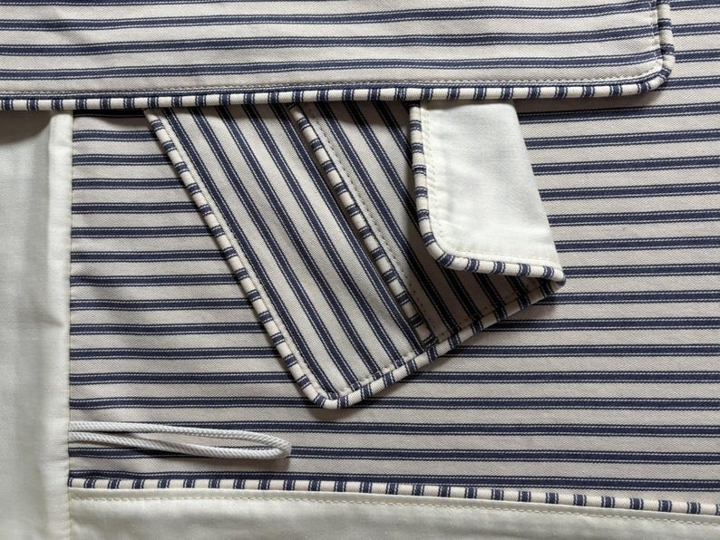 K&ouml;nnte beinhalten: Nahaufnahme eines Stoffes mit blau-wei&szlig;em Streifenmuster. Der Stoff hat eine Tasche mit wei&szlig;em Rand und einem wei&szlig;en Kordelzug. Das Gesamtdesign erinnert an ein klassisches nautisches Thema.
