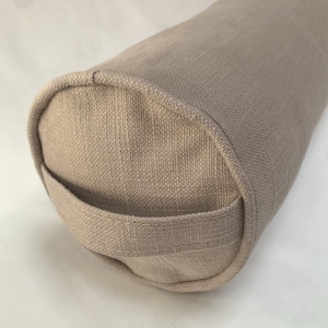 May include: A beige cylindrical bolster pillow with a handle. The pillow is made of a textured fabric and has rounded ends. The handle is a loop of the same fabric, sewn onto the end of the pillow.