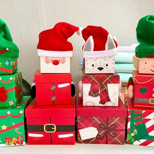 May include: Four stacks of decorative Christmas gift boxes. The first stack is green with a felt elf hat. The second is red with a Santa face and hat. The third is a white dog with a red hat and scarf. The fourth is an elf with a green hat.