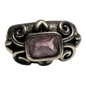 Pode incluir: Um anel de prata com uma pedra preciosa retangular facetada de cor rosa claro no centro. A banda é decorada com volutas intrincadas em relevo e padrões metálicos em espiral que rodeiam a pedra nos lados esquerdo e direito.