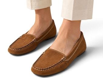 May include: A pair of brown suede loafers featuring a rounded toe, a low heel, and visible white contrast stitching along the moccasin-style seam. The shoes are worn with light-colored trousers, showcasing a simple slip-on design.