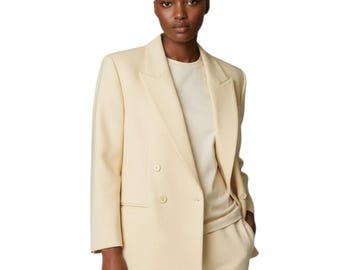 May include: A light cream-colored double-breasted blazer featuring wide lapels and two visible buttons on the front. The blazer is worn over a matching light cream-colored top and trousers, creating a monochromatic ensemble.