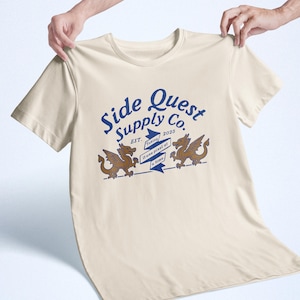 May include: Cream-colored t-shirt featuring the text "Side Quest Supply Co." in blue, with two brown dragon illustrations. The shirt also includes the text "Taking it one pixel at a time."