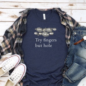 May include: A navy blue t-shirt with a white graphic of a hand with fingers outstretched and the text "Try fingers but hole". The shirt is laid out on a white wooden surface with a plaid shirt, jeans, and white sneakers.