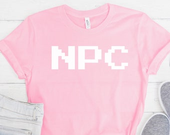May include: A pink t-shirt with the word "NPC" in white pixelated letters. The shirt is laid out on a white wooden surface with a pair of blue denim shorts, a pair of grey trainers, and a pair of sunglasses.