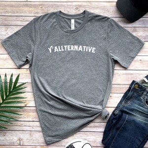 Op de afbeelding: Een grijs t-shirt met de witte tekst "Y'ALLTERNATIVE". Het shirt wordt gecombineerd met een blauwe spijkerbroek, een zwarte pet en sneakers op een houten ondergrond. Het is een casual kledingstuk.