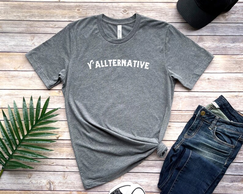 Op de afbeelding: Een grijs t-shirt met de witte tekst "Y'ALLTERNATIVE". Het shirt wordt gecombineerd met een blauwe spijkerbroek, een zwarte pet en sneakers op een houten ondergrond. Het is een casual kledingstuk.