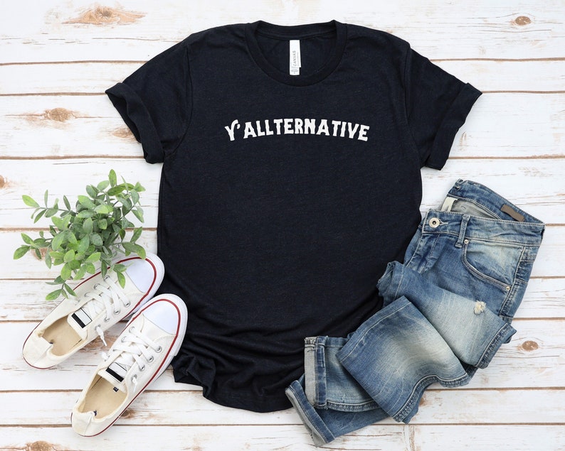 Op de afbeelding: Een donker marineblauw t-shirt met de tekst "Y'ALLTERNATIVE" in het wit. Het shirt wordt gecombineerd met een blauwe jeans en witte sneakers met rode rand. De items zijn gerangschikt op een licht houtnerfoppervlak.