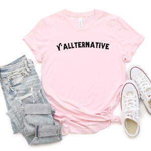 Op de afbeelding: Een lichtroze t-shirt met de tekst "Y'ALLTERNATIVE" in zwarte letters. De afbeelding toont ook een lichtblauwe jeans en witte canvas sneakers.