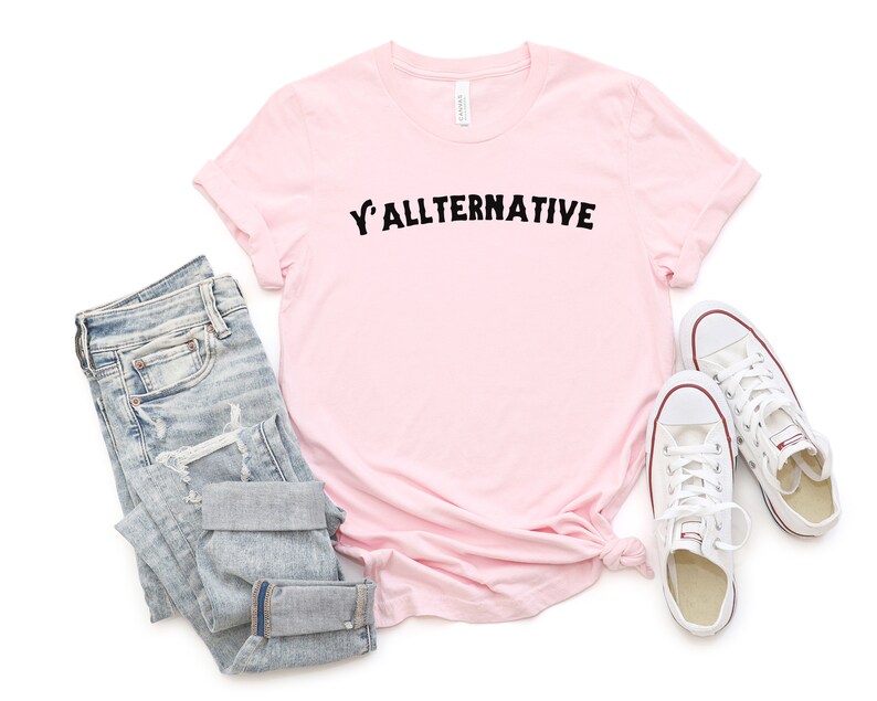 Op de afbeelding: Een lichtroze t-shirt met de tekst "Y'ALLTERNATIVE" in zwarte letters. De afbeelding toont ook een lichtblauwe jeans en witte canvas sneakers.