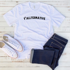 Op de afbeelding: Een wit t-shirt met de tekst "Y'ALLTERNATIVE" in zwart, gecombineerd met een donkerblauwe jeans en witte canvas sneakers. De kleding is geplaatst op een witte houten ondergrond, wat een casual stijl suggereert.