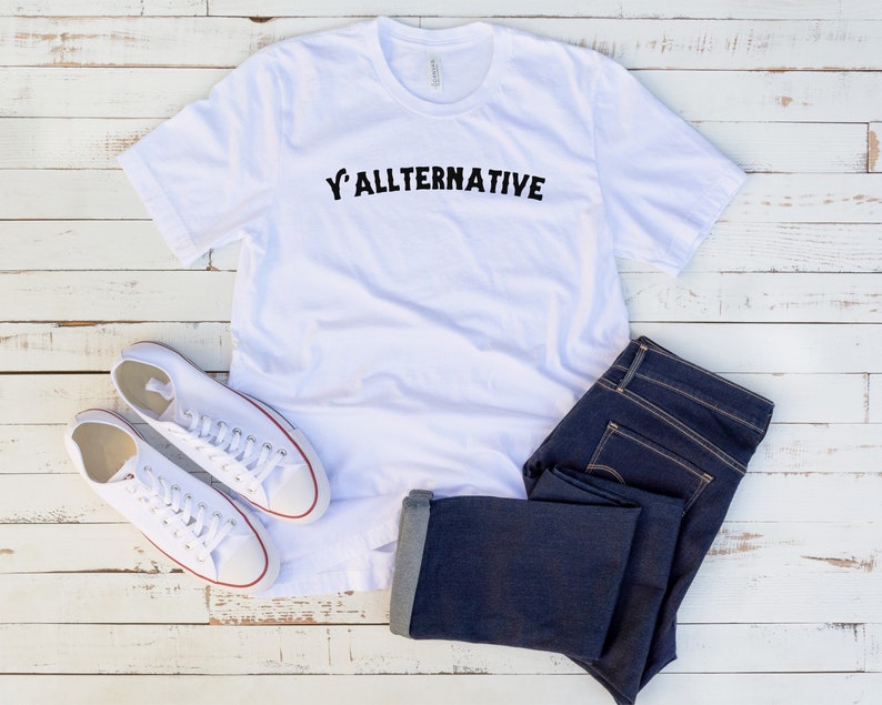 Op de afbeelding: Een wit t-shirt met de tekst "Y'ALLTERNATIVE" in zwart, gecombineerd met een donkerblauwe jeans en witte canvas sneakers. De kleding is geplaatst op een witte houten ondergrond, wat een casual stijl suggereert.