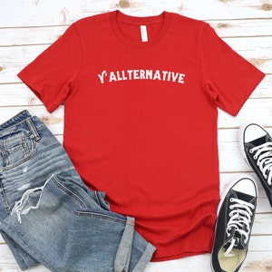 Op de afbeelding: Een rood t-shirt met de witte tekst "Y'allternative", gecombineerd met een blauwe jeans en zwarte canvas sneakers. Het shirt is effen van kleur, met de tekst gecentreerd op de borst.
