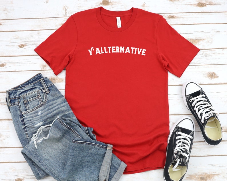 Op de afbeelding: Een rood t-shirt met de witte tekst "Y'allternative", gecombineerd met een blauwe jeans en zwarte canvas sneakers. Het shirt is effen van kleur, met de tekst gecentreerd op de borst.